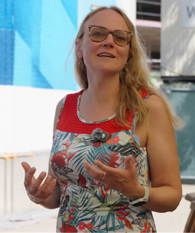 Een vrouw met lang blond haar en een bril glimlacht; ze draagt een kleurrijke bloemenjurk en een rode top. Ze gebaart met haar handen terwijl ze buiten spreekt op een zonnige plek, met achter haar een blauwe muurschildering en een gebouw.