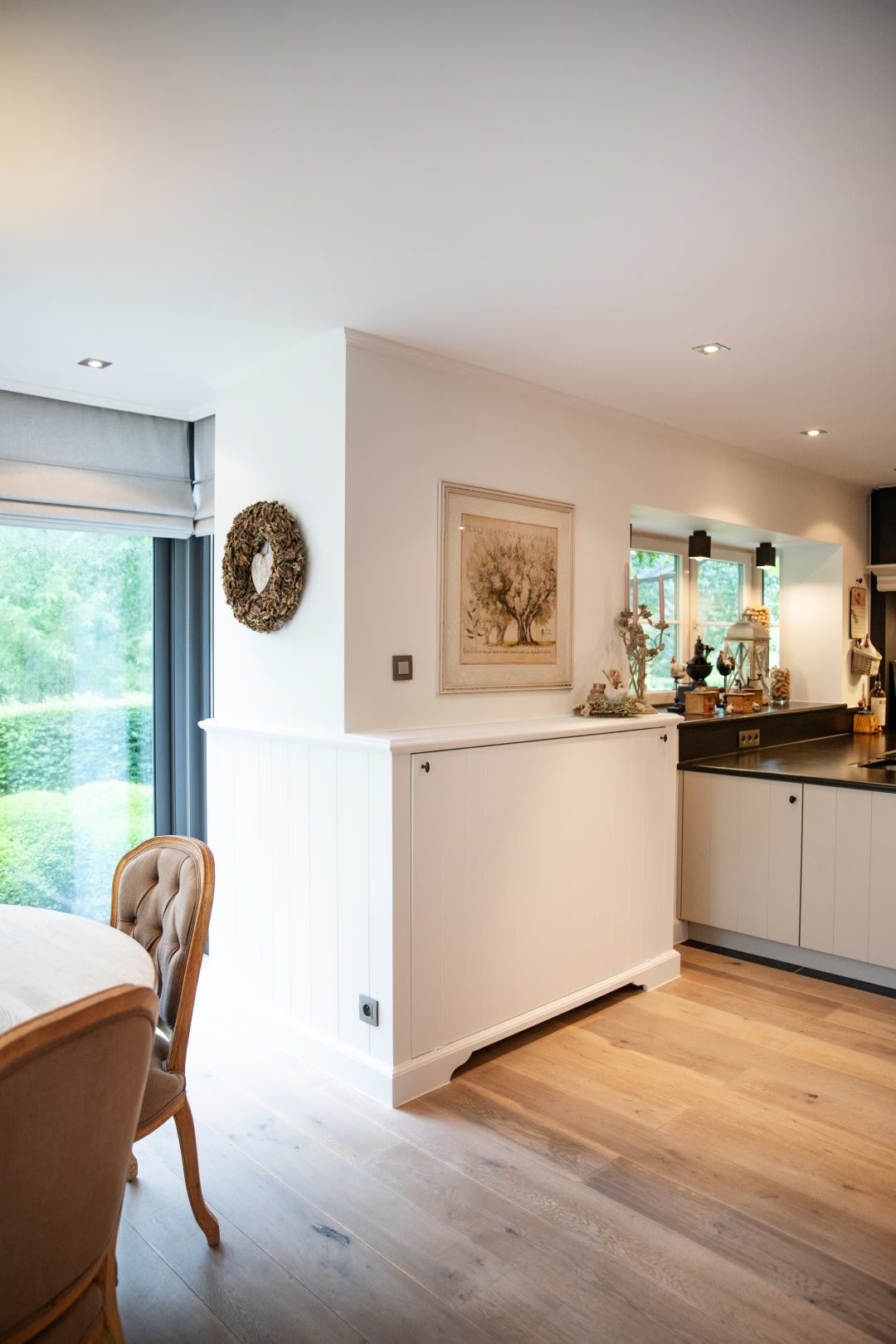 Lichte keuken en eethoek met lichte houten vloeren, wit gelambriseerde muren, groot raam en neutrale inrichting inclusief ingelijste kunst en een decoratieve krans.