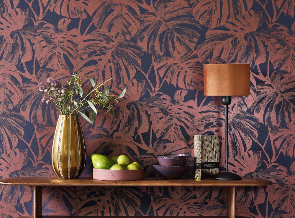 Papier peint avec des feuilles bleu foncé et terracotta  Vase doré avec des fleurs  Table en bois foncé  Lampe de table en terracotta  Bol en bois avec des citrons verts et des avocats  Petits bols violet foncé