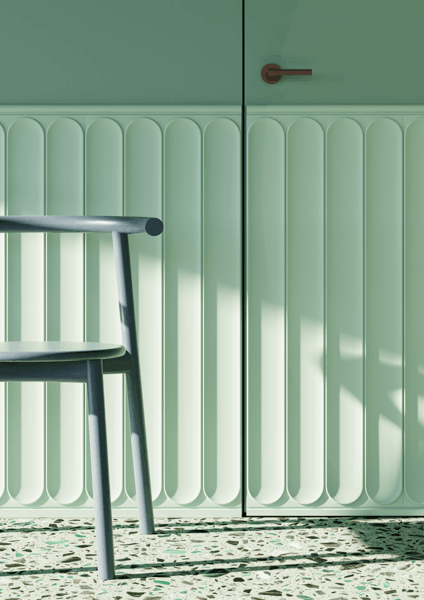 Een lichtgroene muur met verticale lambrisering, een deel van een houten stoel en terrazzovloeren verlicht door natuurlijk zonlicht.
