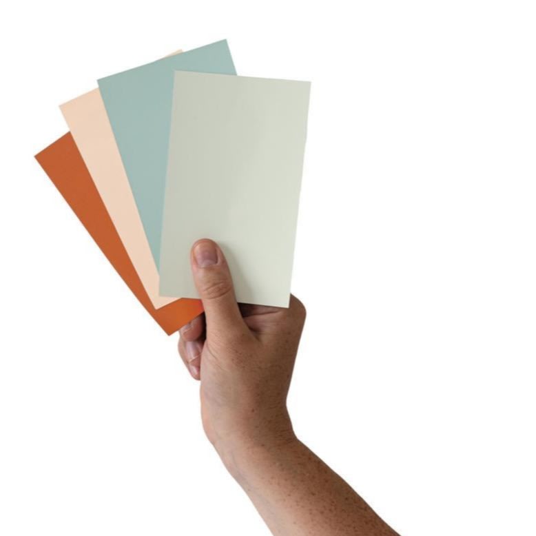 Une main tient quatre cartes avec des échantillons de peinture de différentes couleurs : rouille, pêche, bleu clair et blanc cassé, sur un fond blanc.