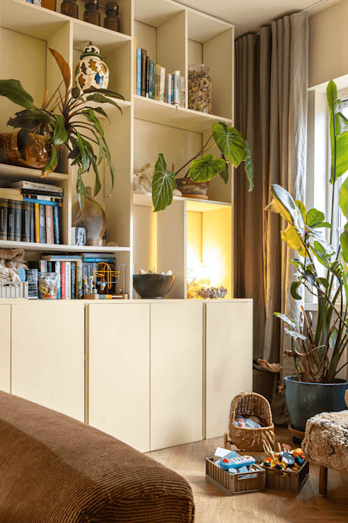 Een gezellige woonkamer met planken met boeken, planten en decoratieve voorwerpen. Speelgoed in manden ligt op de houten vloer bij een bruine bank en een raam met gordijnen.