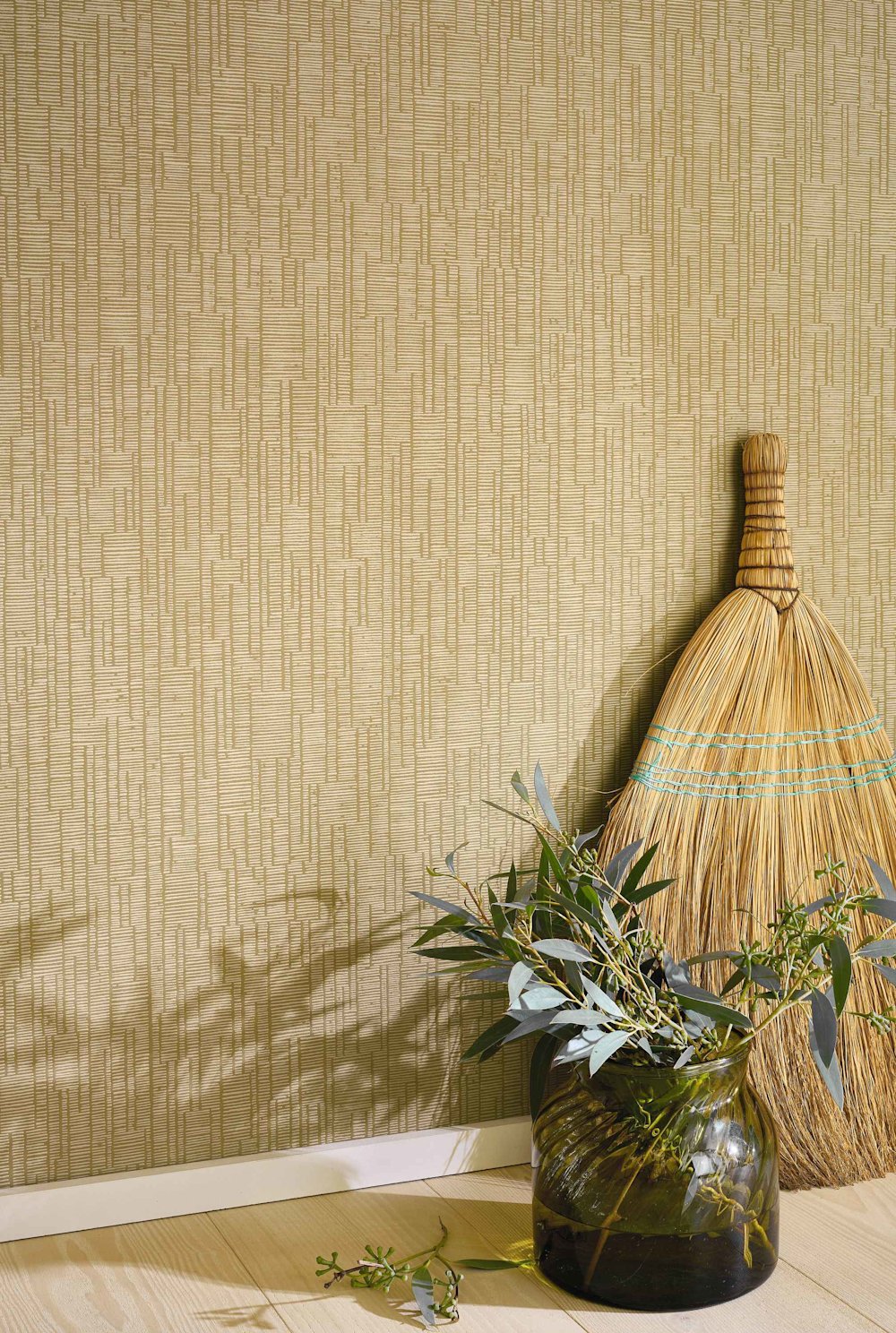 Papier peint texturé couleur sable  Balai en osier avec accents bleus  Vase en verre vert avec eucalyptus  Parquet clair  Plinthes blanches