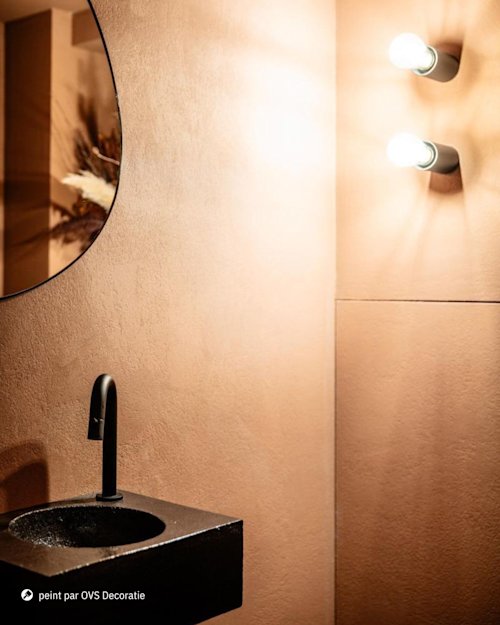 Une salle de bains moderne avec un miroir rond, un robinet et un lavabo noirs, des murs beiges et deux ampoules montées verticalement.