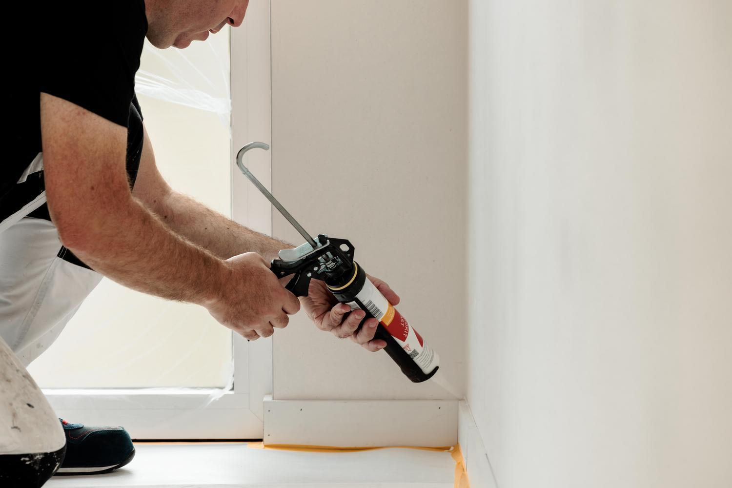 Een persoon brengt kit aan langs de onderkant van een witte muur met een kitpistool. Hij knielt op de grond en draagt een witte broek met zwarte vlekken en een zwart overhemd.