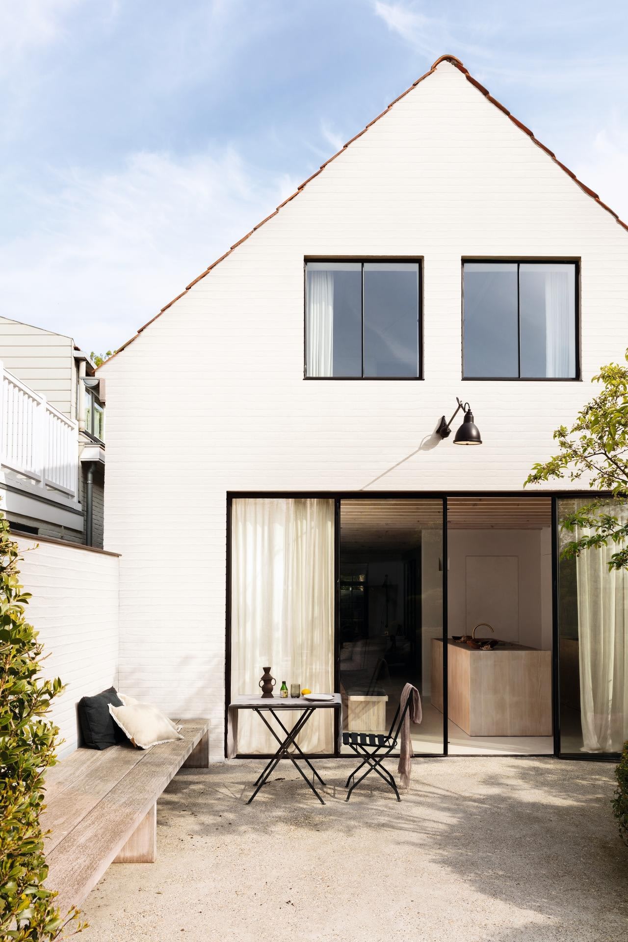 Een modern wit huis met grote zwarte ramen, een schuin dak en een patio met een kleine tafel, twee stoelen en een ingebouwde houten bank omringd door groen op een zonnige dag.