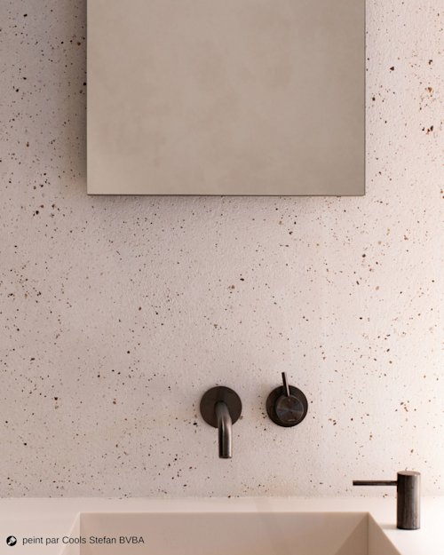 Mur de salle de bain minimaliste avec un grand miroir, un robinet noir et un distributeur de savon au-dessus d'un lavabo blanc, avec une finition texturée et mouchetée.