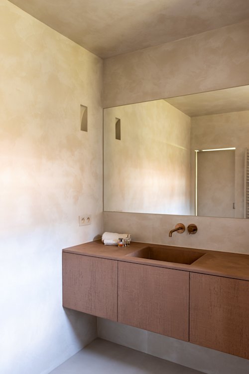 Une salle de bain minimaliste avec un grand miroir, une vanité flottante marron, un lavabo intégré, un robinet mural et des murs aux tons neutres.