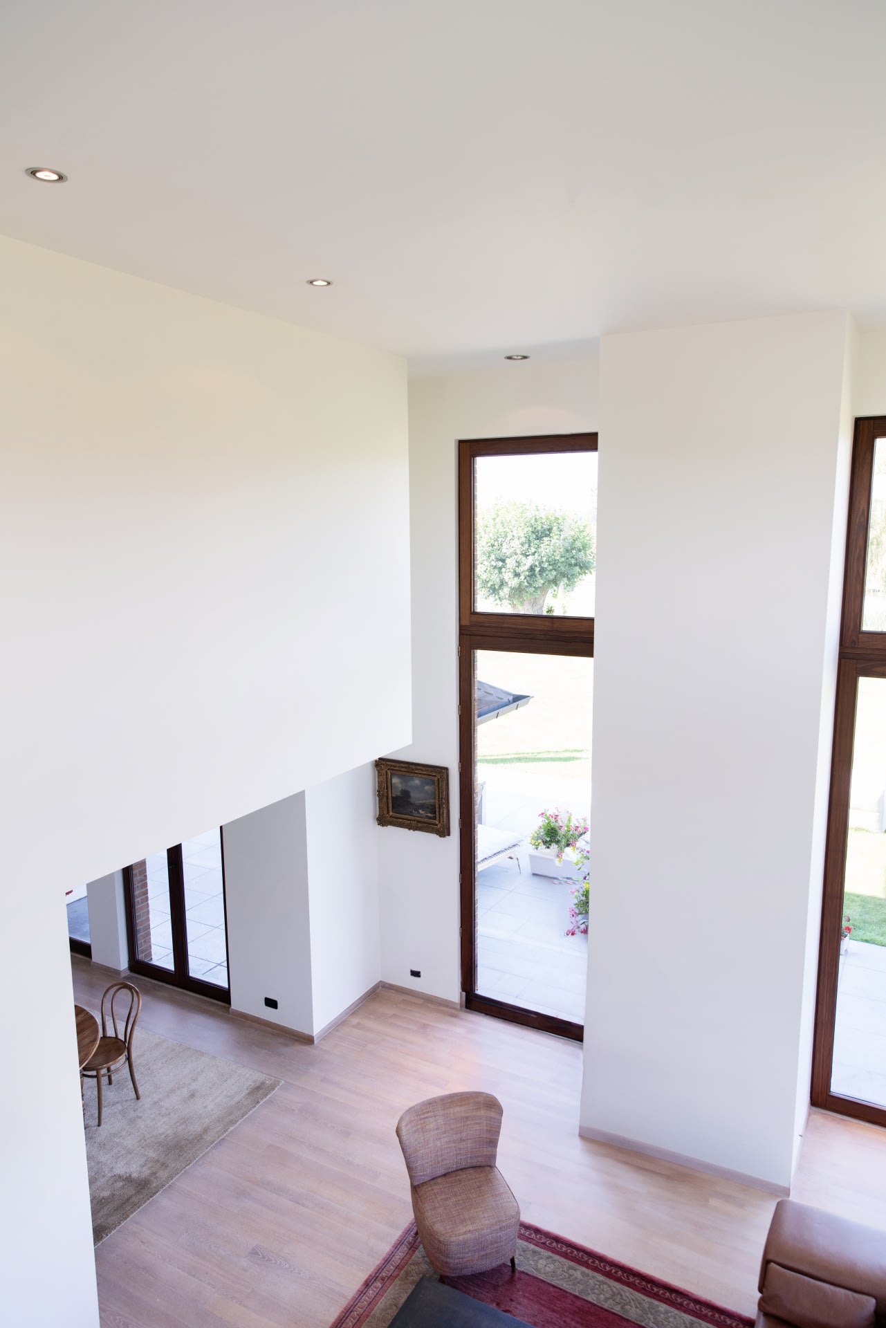 Salon moderne avec de hauts plafonds, de grandes fenêtres, du parquet clair et un mobilier minimaliste comprenant une chaise et un tapis, vu depuis un angle surélevé.