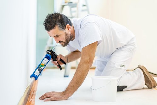 Een man in witte werkkleding knielt op de grond en brengt kit aan langs de onderkant van een muur met een kitpistool; een witte emmer staat vlakbij.