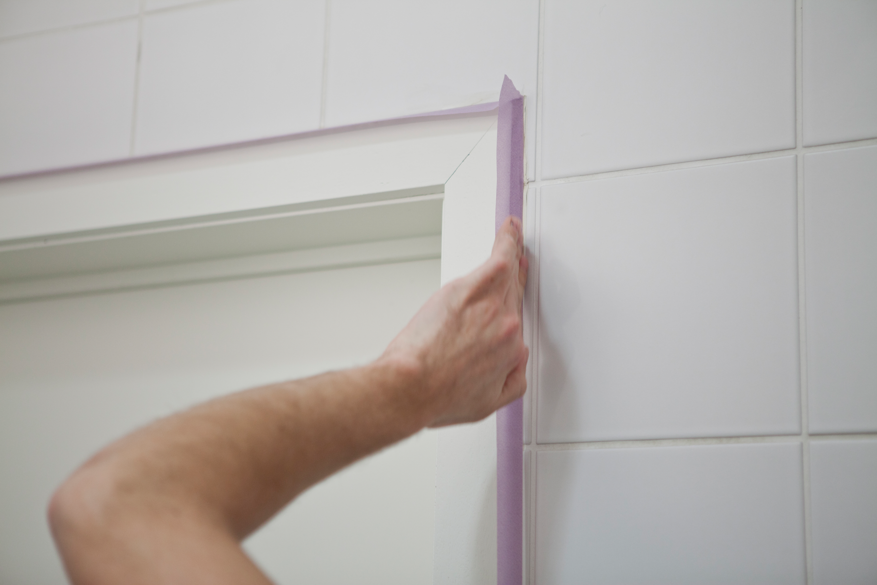 La main d’une personne applique du ruban de masquage violet le long du bord d’un encadrement de porte blanc, à côté d’un mur carrelé, afin de préparer la zone pour la peinture ou le jointoiement.