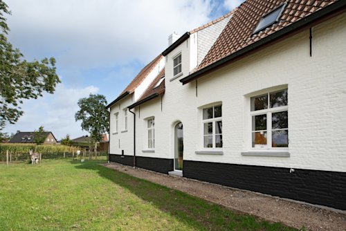 Een wit bakstenen huis met een bruin pannendak staat op een grasveld met bomen en een ezel zichtbaar op de achtergrond.