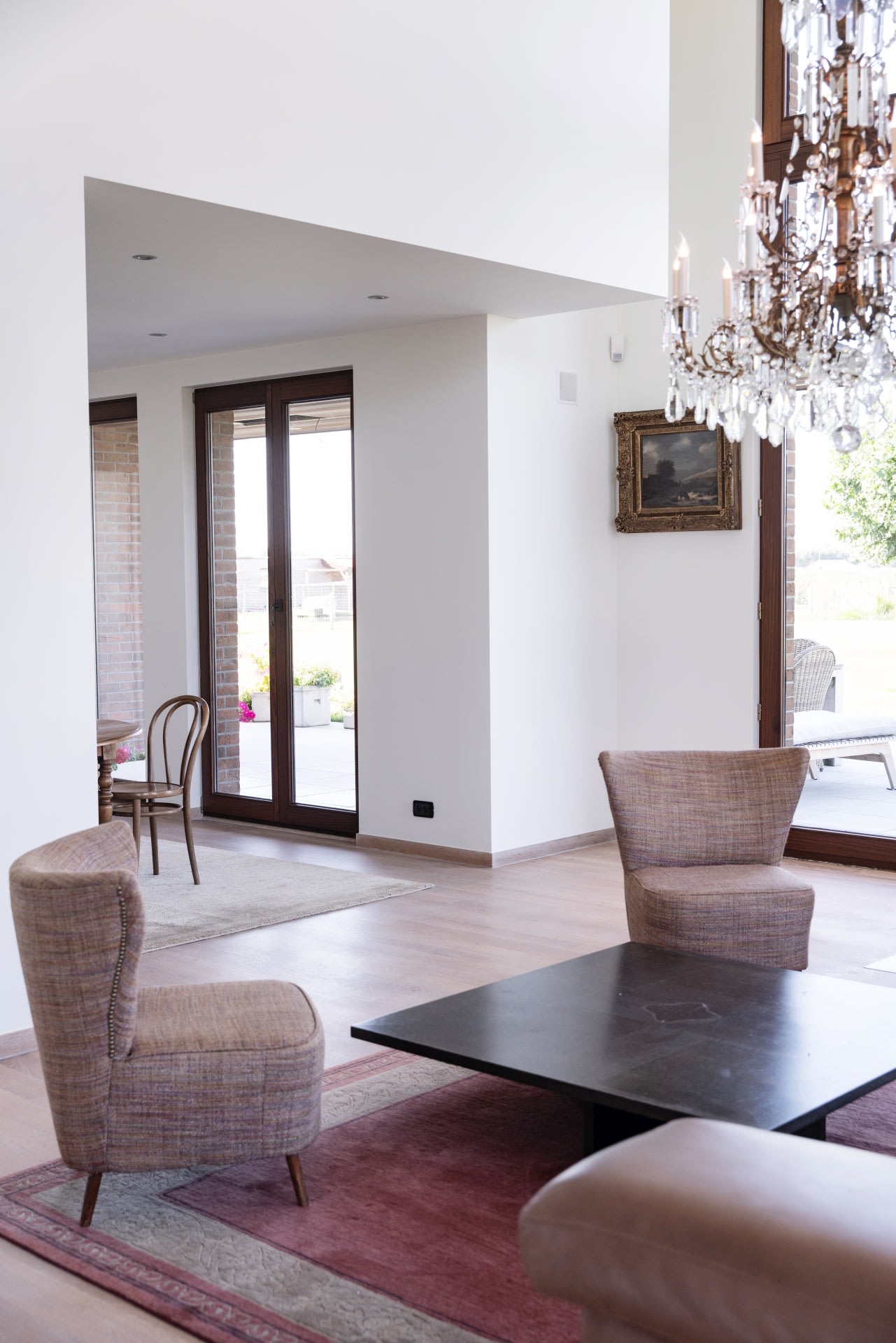 Un salon moderne avec des murs clairs, de grandes fenêtres, deux fauteuils rembourrés, une table basse noire, un tapis, un lustre et un tableau encadré accroché au mur.