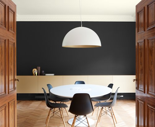 Een moderne eetkamer met een ronde witte tafel, zes zwarte stoelen, een grote koepelvormige hanglamp en lichthouten kasten tegen een zwarte accentmuur.
