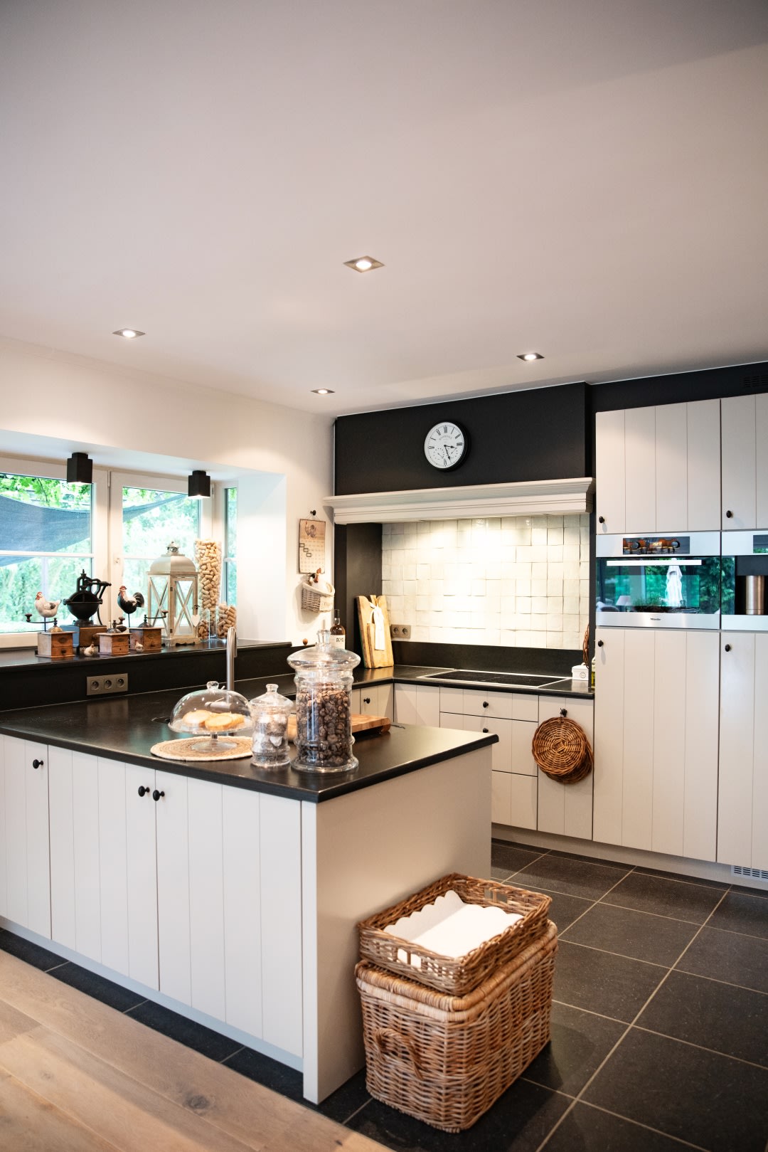 Moderne keuken met witte kasten, zwarte werkbladen, een centraal eiland, inbouwapparatuur, een wandklok en een rieten mand op de vloer.