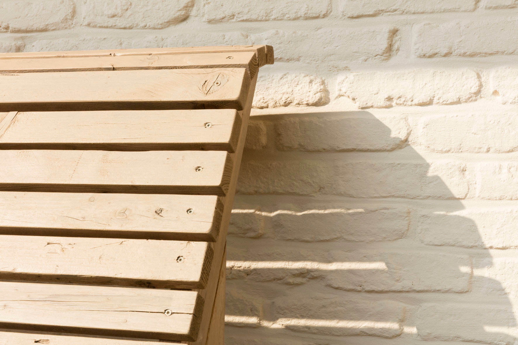 Houten latten die in een diagonaal patroon zijn aangebracht en met zichtbare schroeven zijn bevestigd, werpen in het zonlicht een schaduw op een witte bakstenen muur.