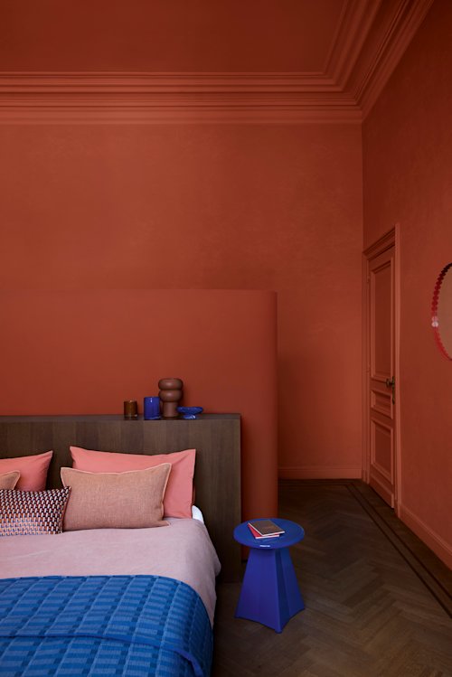 Une chambre avec des murs terracotta, un lit en bois avec des coussins roses et beiges, une couverture bleue et une petite table d’appoint bleue sur laquelle est posé un carnet rouge. Le sol est en parquet à chevrons.