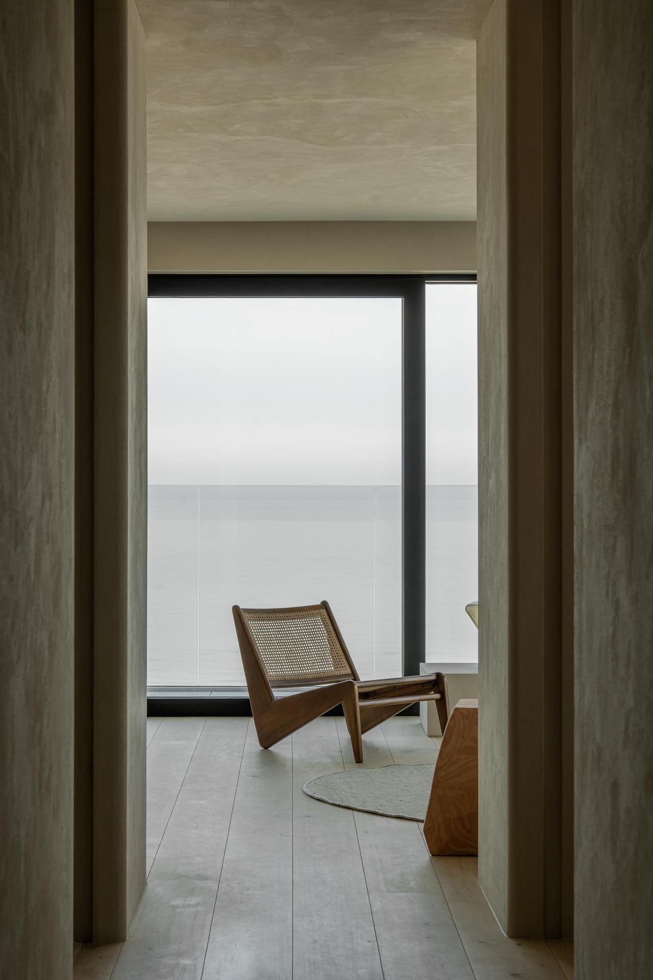 Een minimalistische kamer met lichte houten vloeren en muren, met een geweven loungestoel en klein tafeltje bij een groot raam dat uitkijkt op een kalm, wazig zeegezicht.