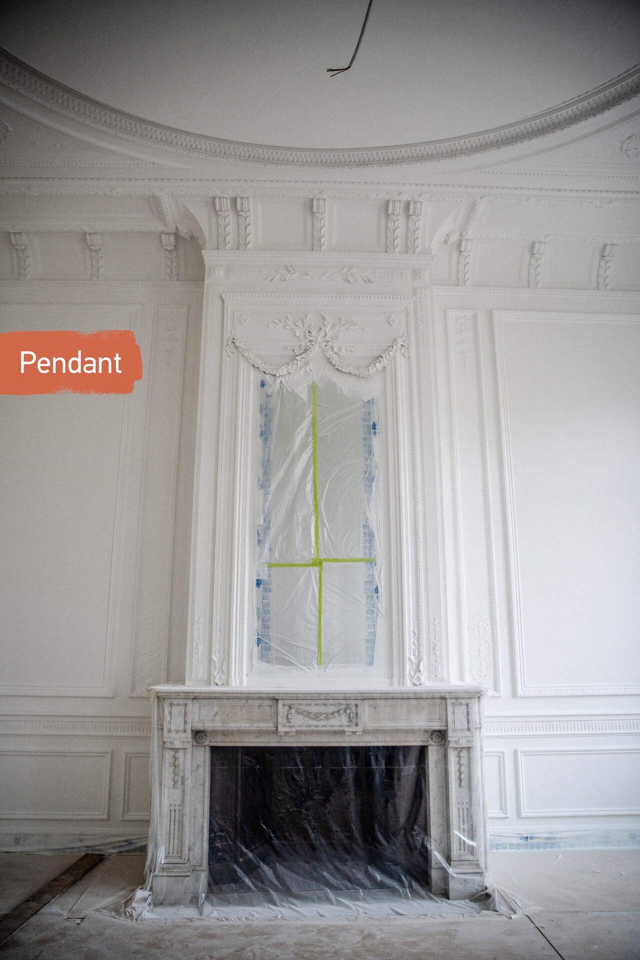 Une cheminée en marbre dans une pièce blanche et ornée ; le manteau et la fenêtre au-dessus sont recouverts d'un film plastique protecteur, signe de rénovations en cours. À gauche, on peut lire « Pendant ».