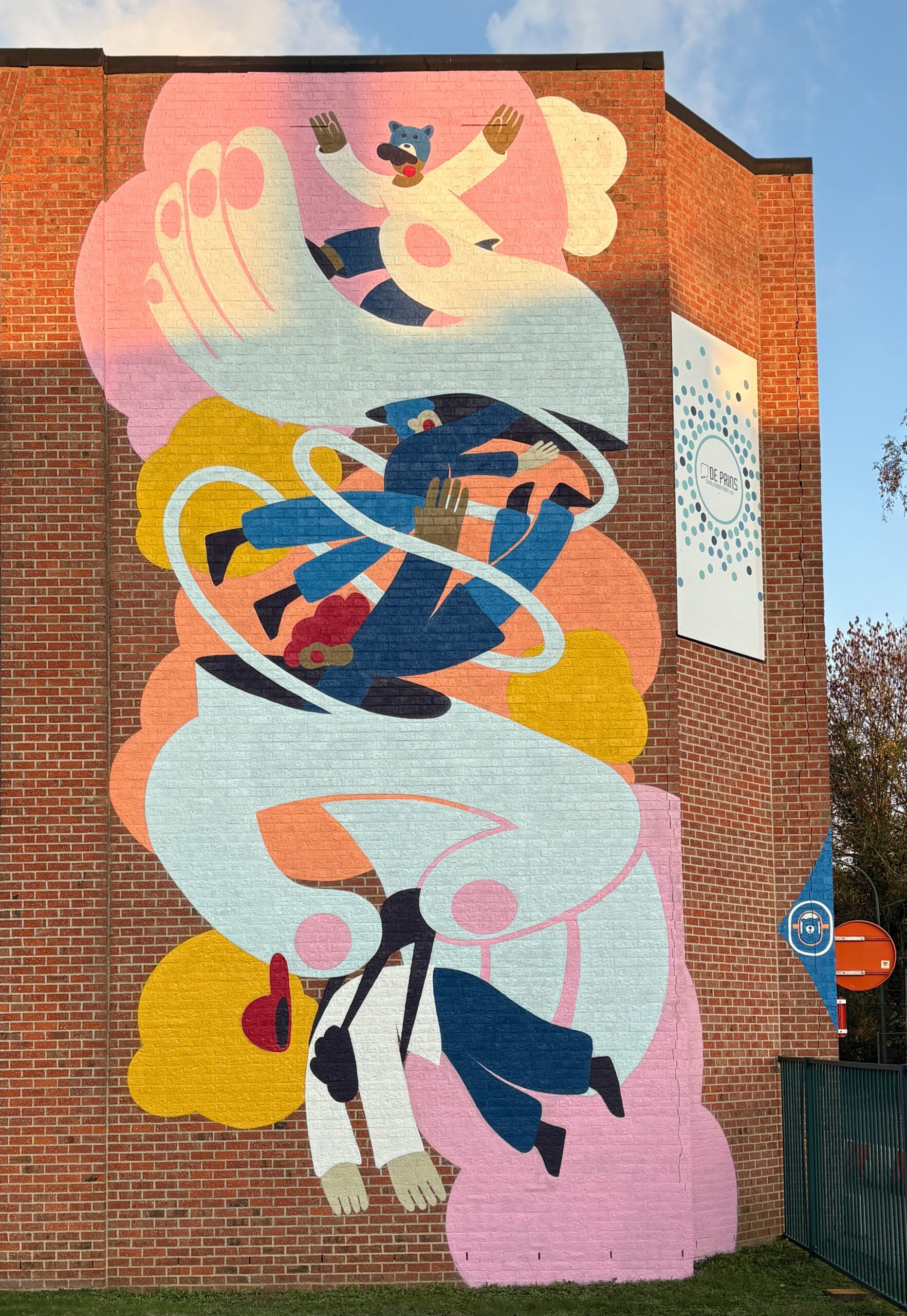 Een kleurrijke muurschildering op een bakstenen muur toont abstracte menselijke figuren in blauw, roze, geel en wit, die lijken te zweven en zich verstrengelen tussen wervelende vormen en wolken, met expressieve gezichten en handen.