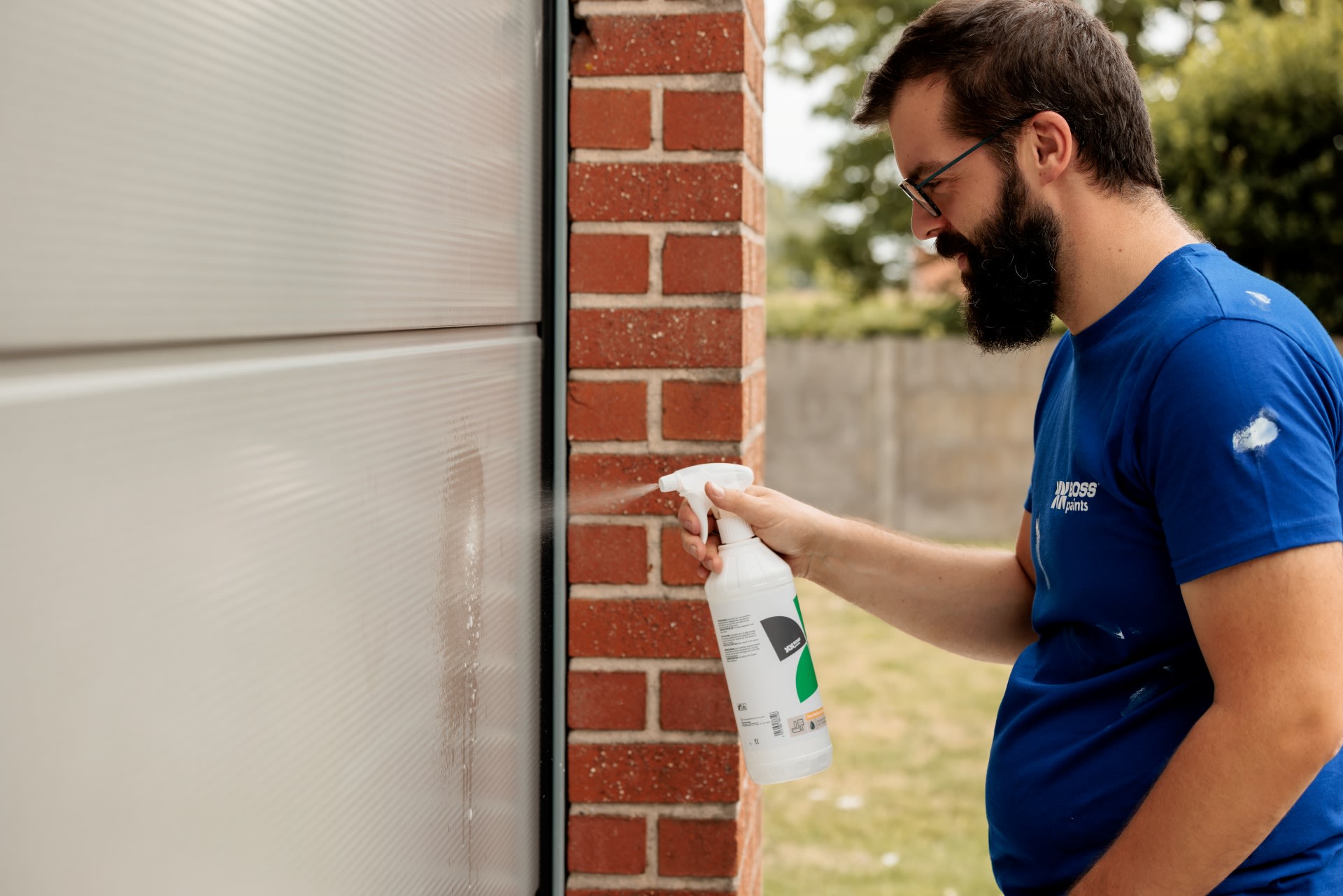 Een man in een blauw shirt spuit schoonmaakmiddel op een witte garagedeur naast een bakstenen muur.