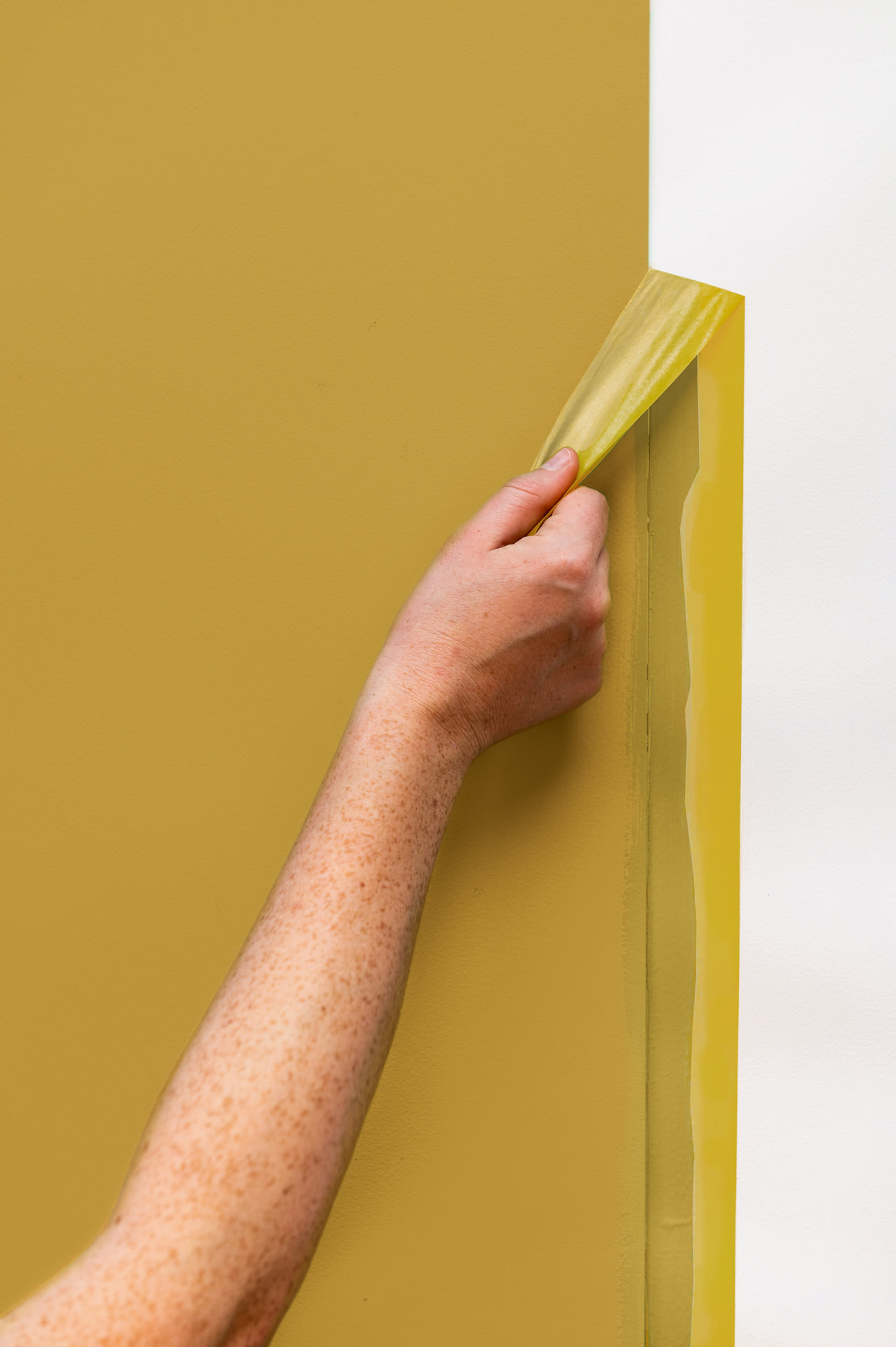 Une main décolle du ruban de masquage jaune du bord d'un mur fraîchement peint en jaune moutarde, faisant apparaître une ligne nette entre la peinture et le mur blanc.