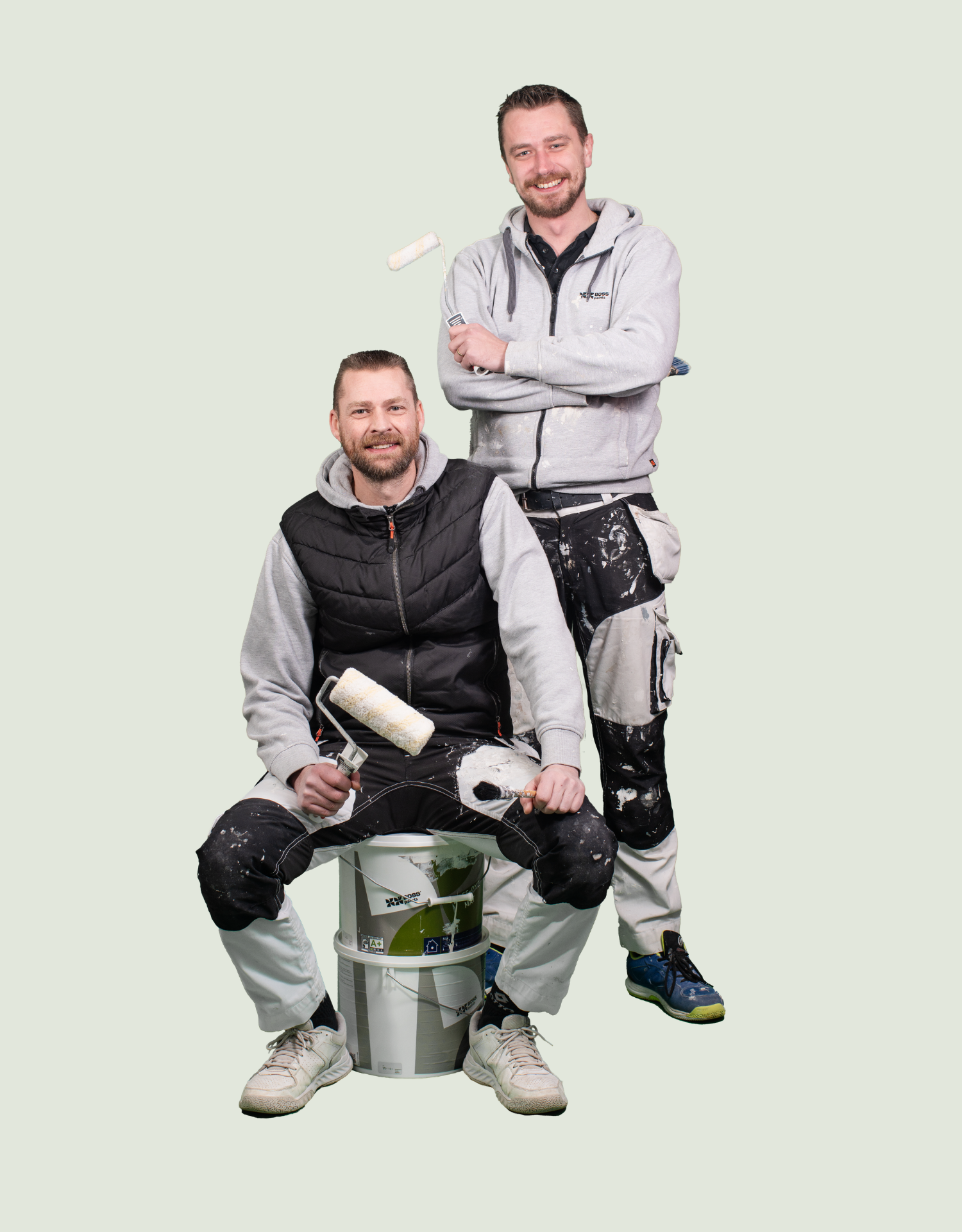 Twee lachende schilders poseren tegen een lichte achtergrond. De ene zit op een stapel verfemmers met een roller en kwast, de andere staat achter hem met de armen over elkaar. Beiden dragen werkkleding die bespat is met verf.