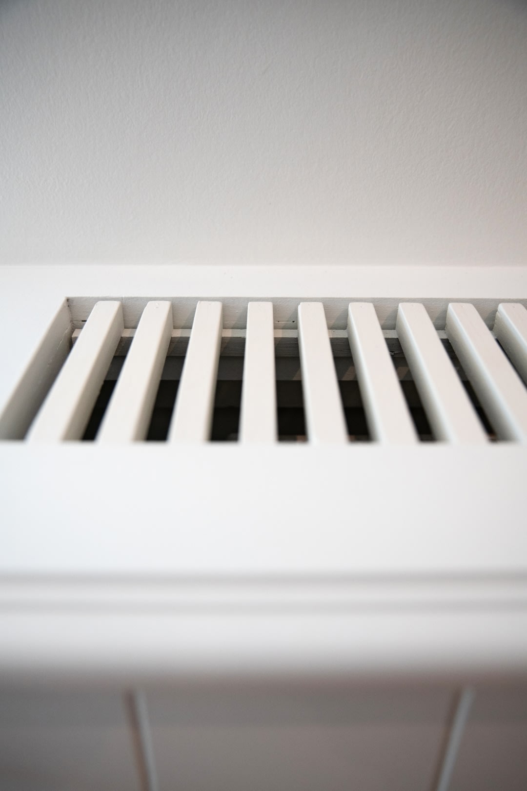 Een close-up van een wit ventilatierooster of registerafdekking met schuine lamellen, gemonteerd op een witte muur.