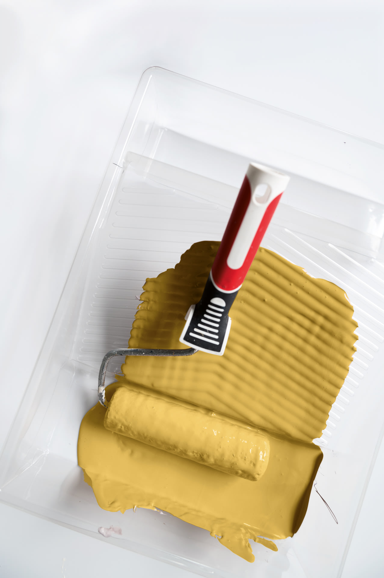 Un rouleau à peinture muni d'un manche rouge et blanc repose dans un bac en plastique transparent rempli de peinture jaune moutarde, prêt à l'emploi.