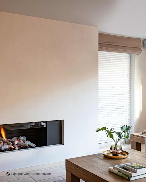 Un salon moderne avec une cheminée encastrée, une table en bois, des plantes en pots sur un plateau et une grande fenêtre avec des stores blancs laissant entrer la lumière naturelle.