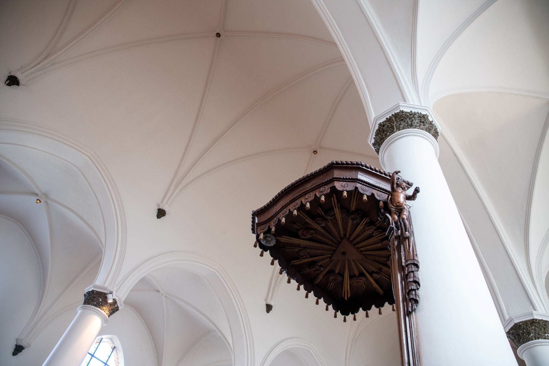 Wit gewelf met decoratieve zuilen en een grote, sierlijke houten baldakijn aan één zuil, van onderaf gezien in een kerk.