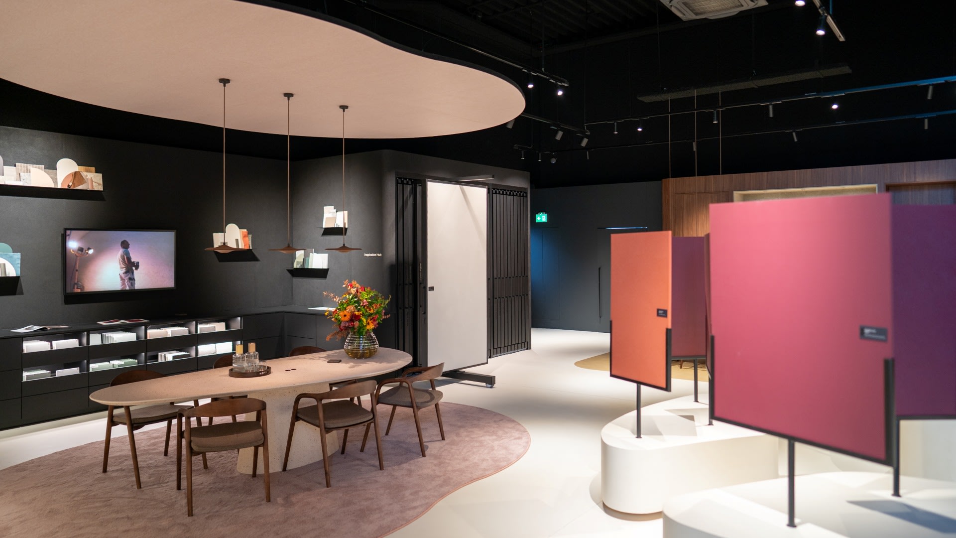 Modern showroominterieur met een gebogen vergadertafel met stoelen, aan de muur bevestigde displayplanken en verschillende vrijstaande rechthoekige privacyschermen in rode en oranje tinten.
