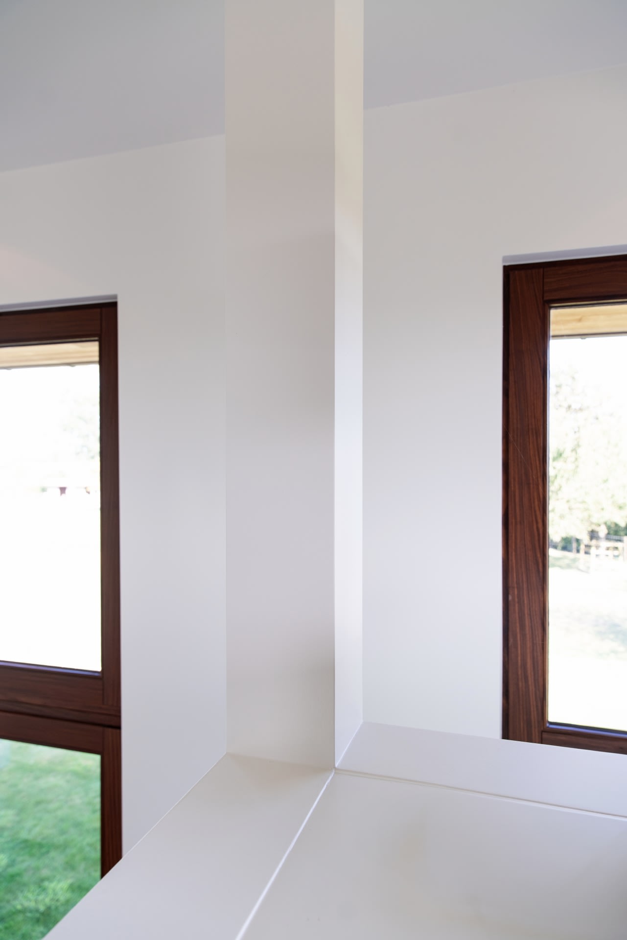 Un mur d'angle blanc avec de la lumière naturelle qui pénètre à l'intérieur par deux fenêtres à encadrements en bois et laisse voir l'herbe verte à l'extérieur.