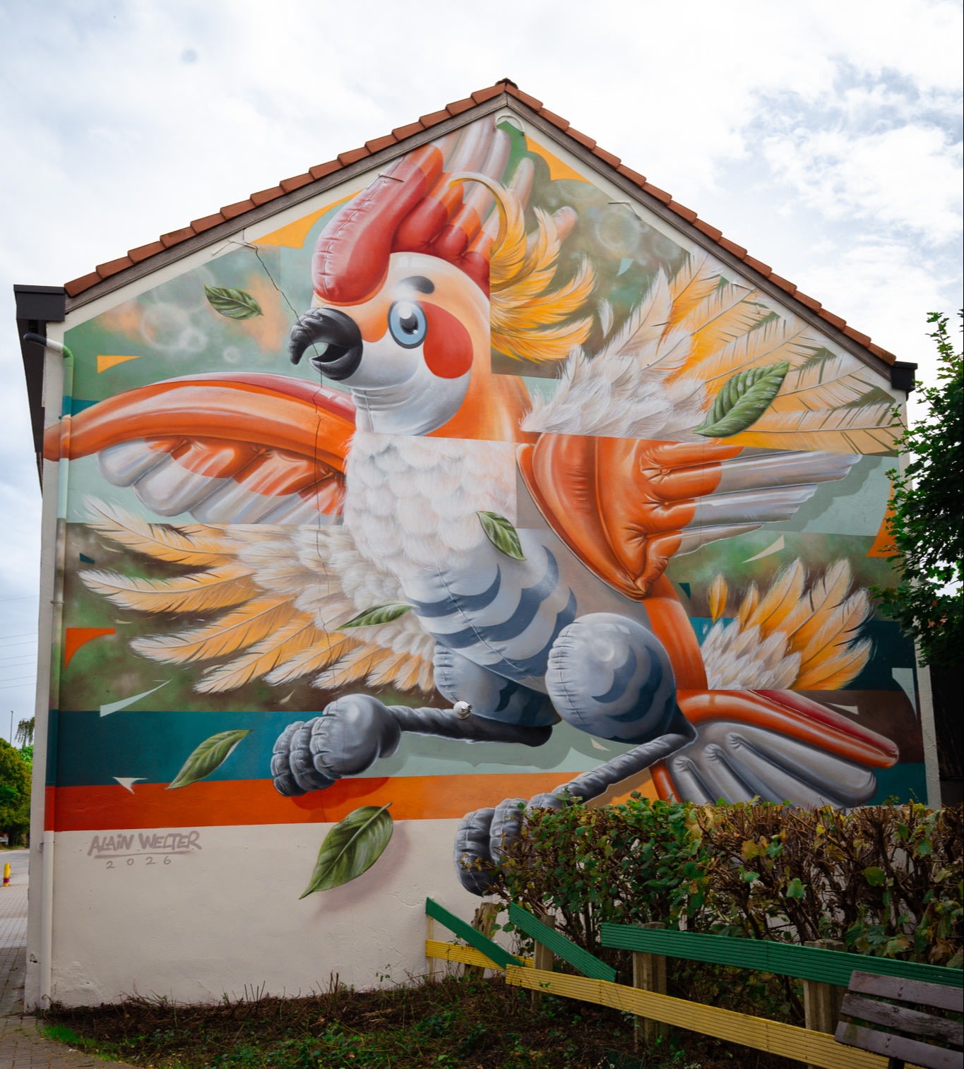Een kleurrijke muurschildering van een grote, gestileerde papegaai met oranje, gele en witte veren siert de zijkant van een huis, met levendige bladeren en dynamische bewegingen.