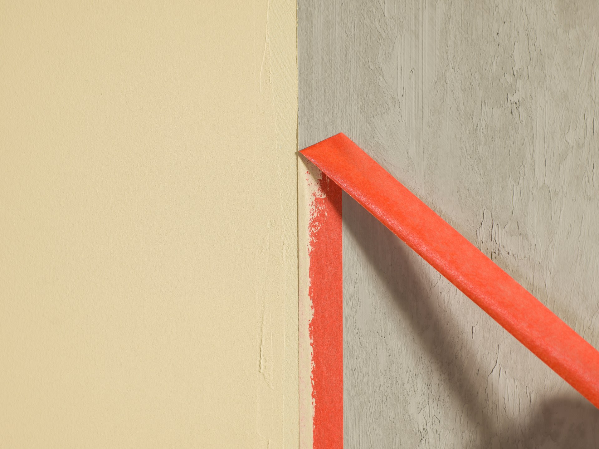 Un morceau de ruban de peintre rouge est retiré du bord où un mur beige et un mur texturé gris se rencontrent.