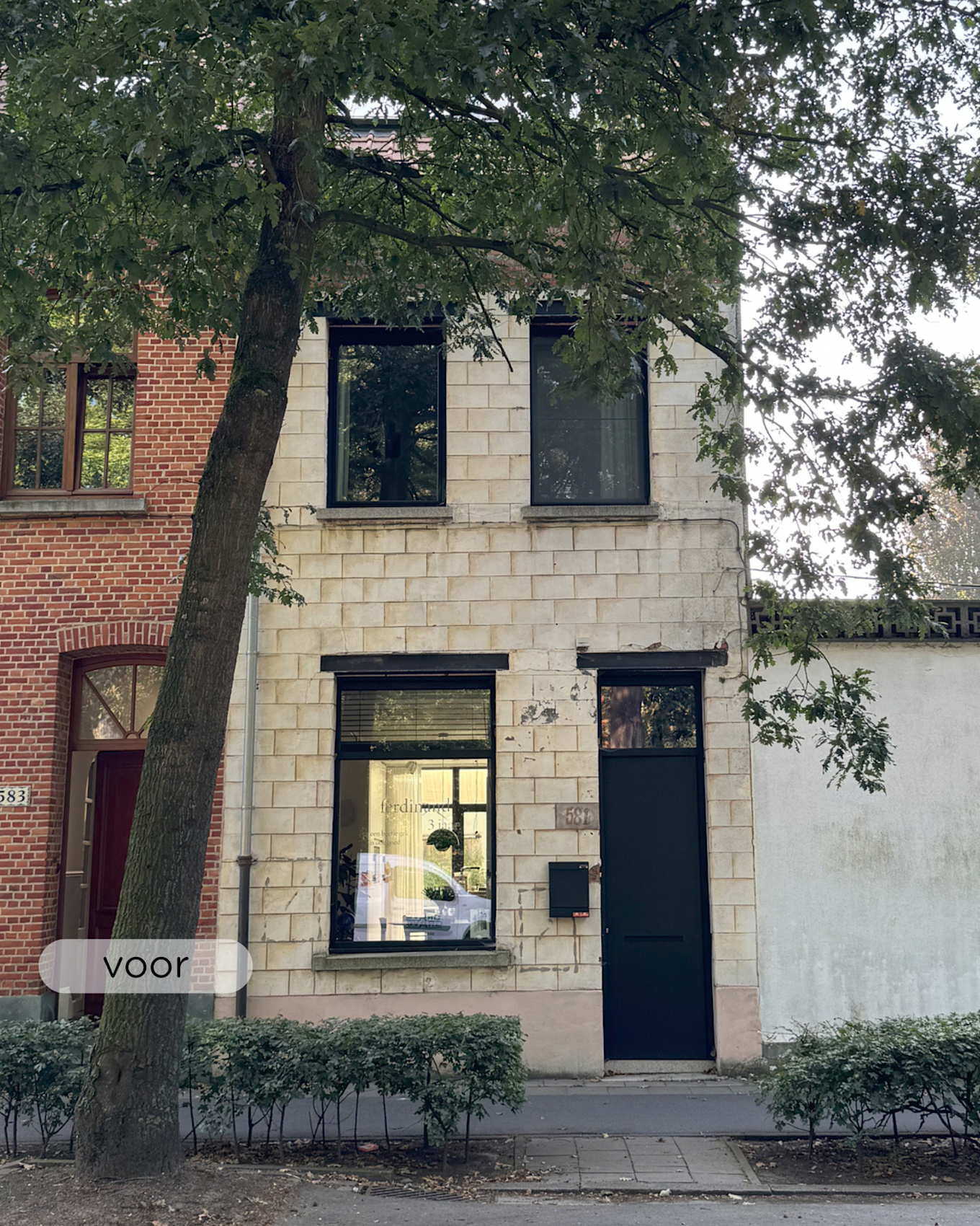 Een klein crèmekleurig gebouw van twee verdiepingen met zwarte deuren en ramen, gedeeltelijk aan het oog onttrokken door een grote boom, staat tussen een rood bakstenen gebouw en een witte muur langs een stadsstraat.