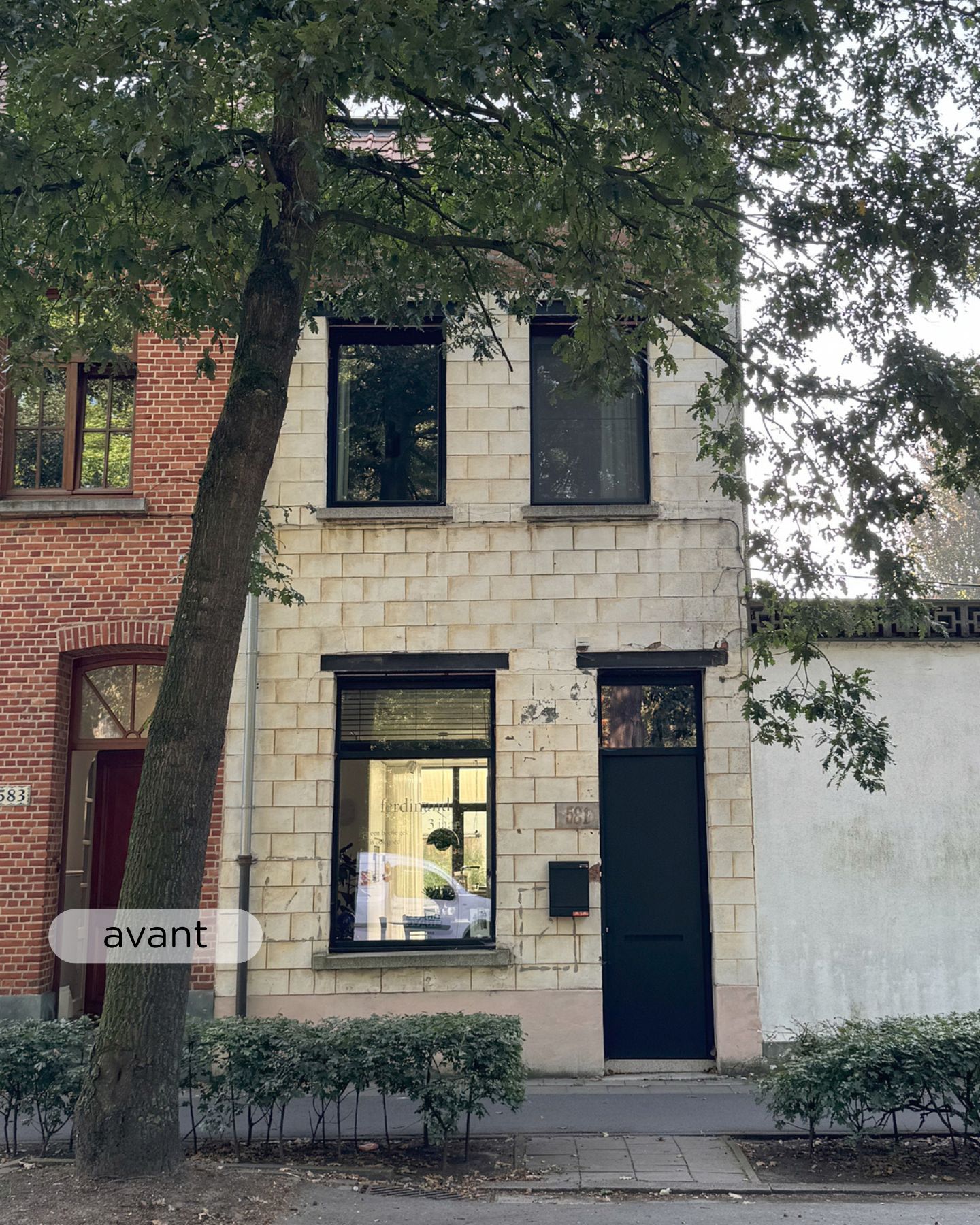 Un petit bâtiment de deux étages, de couleur crème, avec des portes et des fenêtres noires, partiellement masqué par un grand arbre, se dresse entre un bâtiment en briques rouges et un mur blanc le long d'une rue de la ville.
