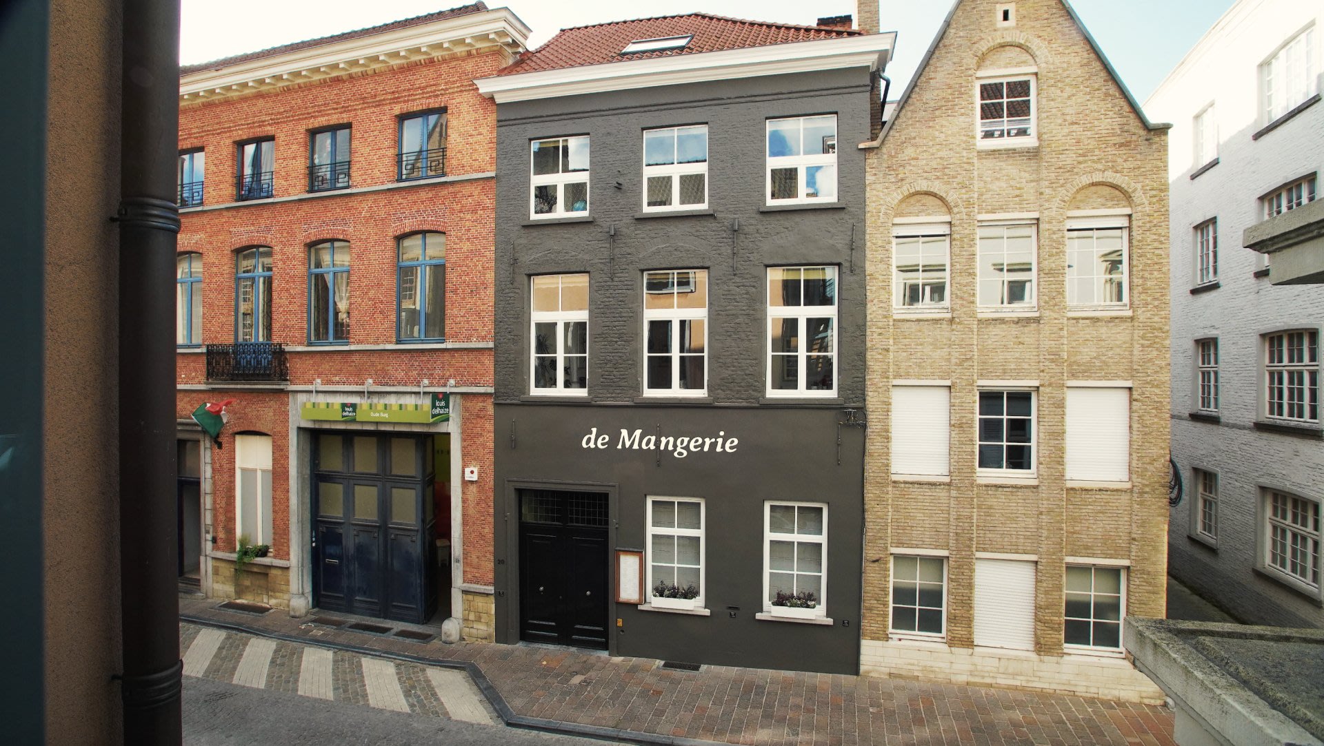 Une rue pavée étroite bordée de trois immeubles à plusieurs étages, dont l'un arbore une enseigne « de Mangerie » sur sa façade gris foncé.