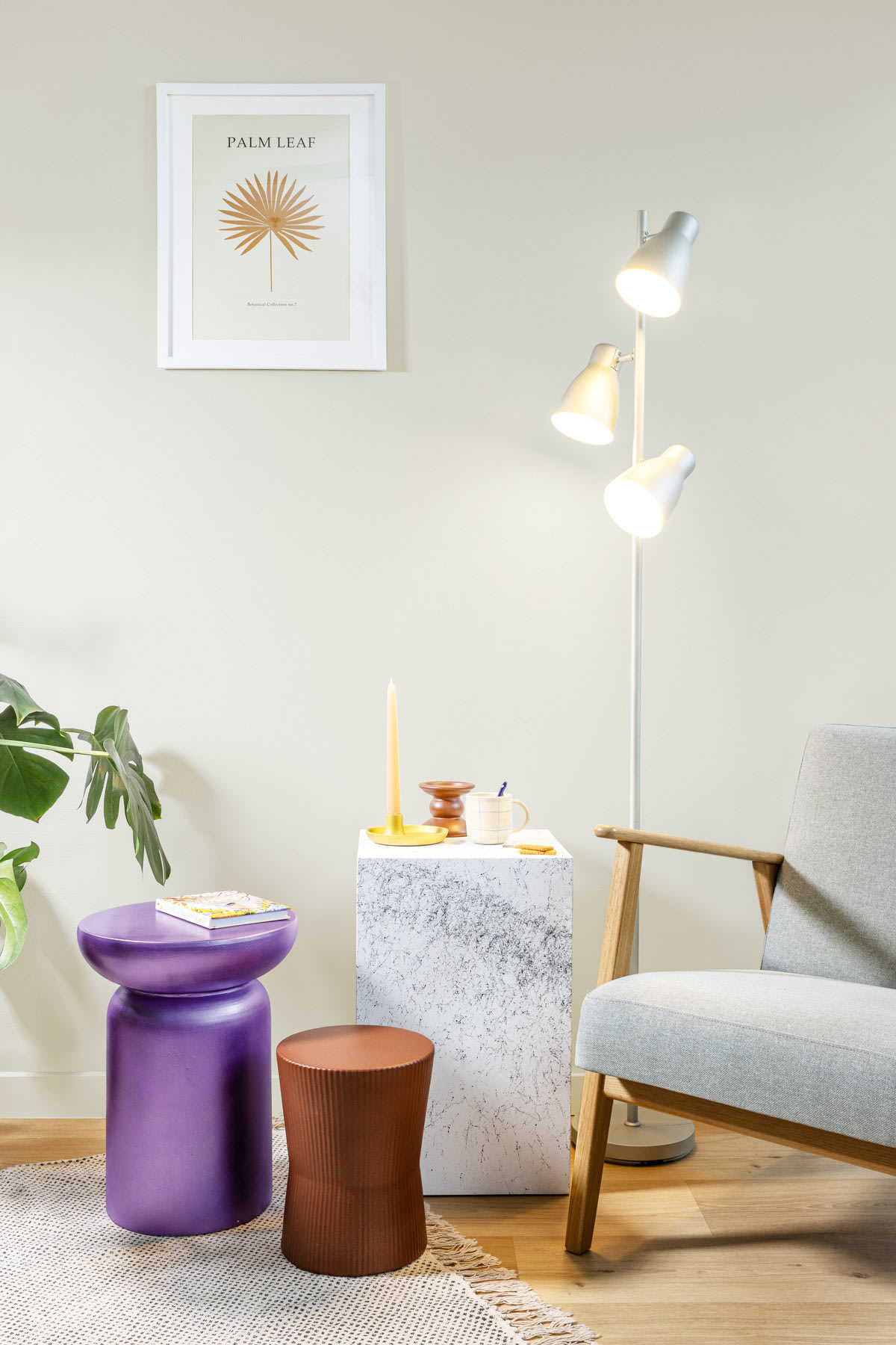 Een moderne woonkamerhoek met een grijze fauteuil, een marmeren bijzettafel, paarse en bruine krukjes, een staande lamp, een ingelijste bladprint en een kamerplant.