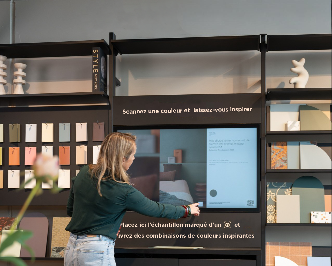 Une femme utilise l'écran tactile d'une borne d'inspiration couleur, entourée d'échantillons de peinture étiquetés « couleurs tendance ». L'écran invite les utilisateurs à scanner une couleur et à découvrir des combinaisons de couleurs assorties.