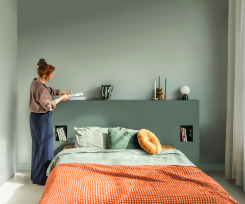 Een vrouw schikt boeken op een plank boven een keurig opgemaakt bed met groen en oranje beddengoed. Nederlandse tekst biedt kleuradvies aan huis met 100% cashback en een knop om een afspraak te maken.
