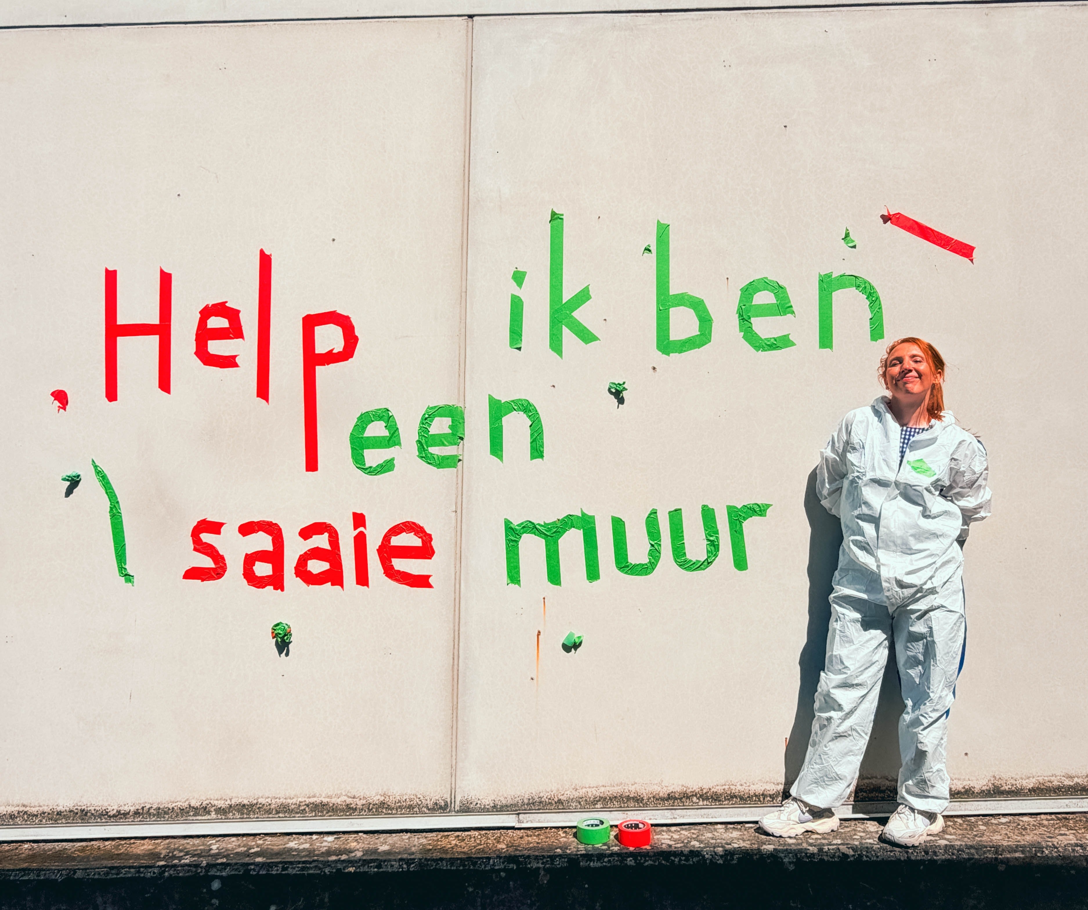 Een persoon in een wit beschermpak staat glimlachend naast een muur met Nederlandse graffiti in rode en groene letters met de tekst "Help, ik ben een saaie muur," wat vertaald kan worden als "Help, ik ben een saaie muur.