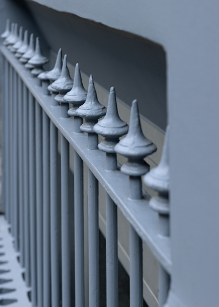 Close-up van een metalen hek met spitse, decoratieve eindstukken, geschilderd in een gedempte grijsblauwe kleur, uitgelijnd voor een bijpassende muur.