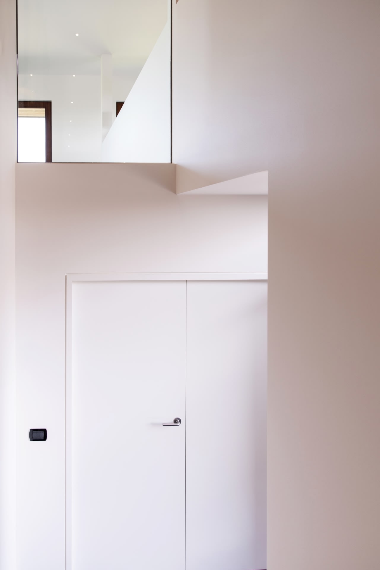 Intérieur minimaliste avec des murs blancs, une porte blanche fermée, une balustrade en verre au-dessus et un interrupteur noir sur le mur gauche. La lumière naturelle pénètre par une fenêtre dans la partie supérieure.