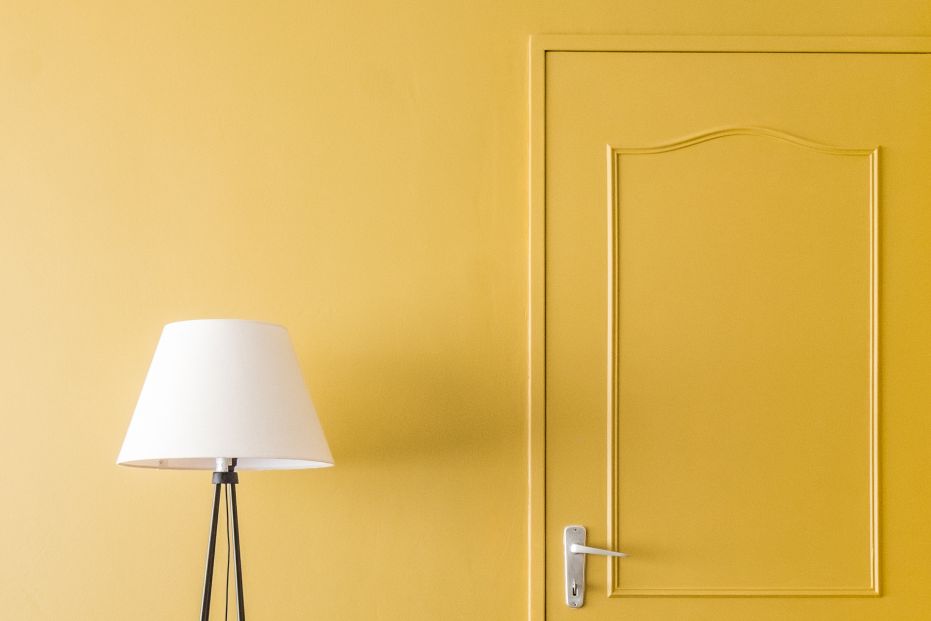Un abat-jour blanc monté sur un trépied noir se trouve à côté d'une porte jaune aux ornements raffinés et se fond harmonieusement dans un mur jaune assorti, au sein d'un intérieur minimaliste et moderne.