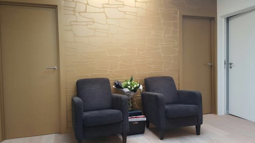 Twee donkergrijze fauteuils met daartussen een zwart tafeltje en een boeket witte bloemen staan tegen een gouden muur met een stenen patroon, geflankeerd door twee gouden deuren.