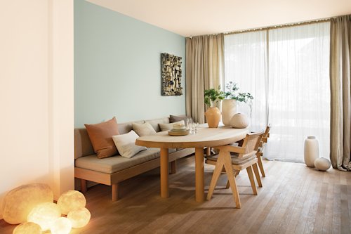 woonkamer met blauwe muur, houten tafel met zetel en houten stoelen, grijze gordijnen en houten vloer