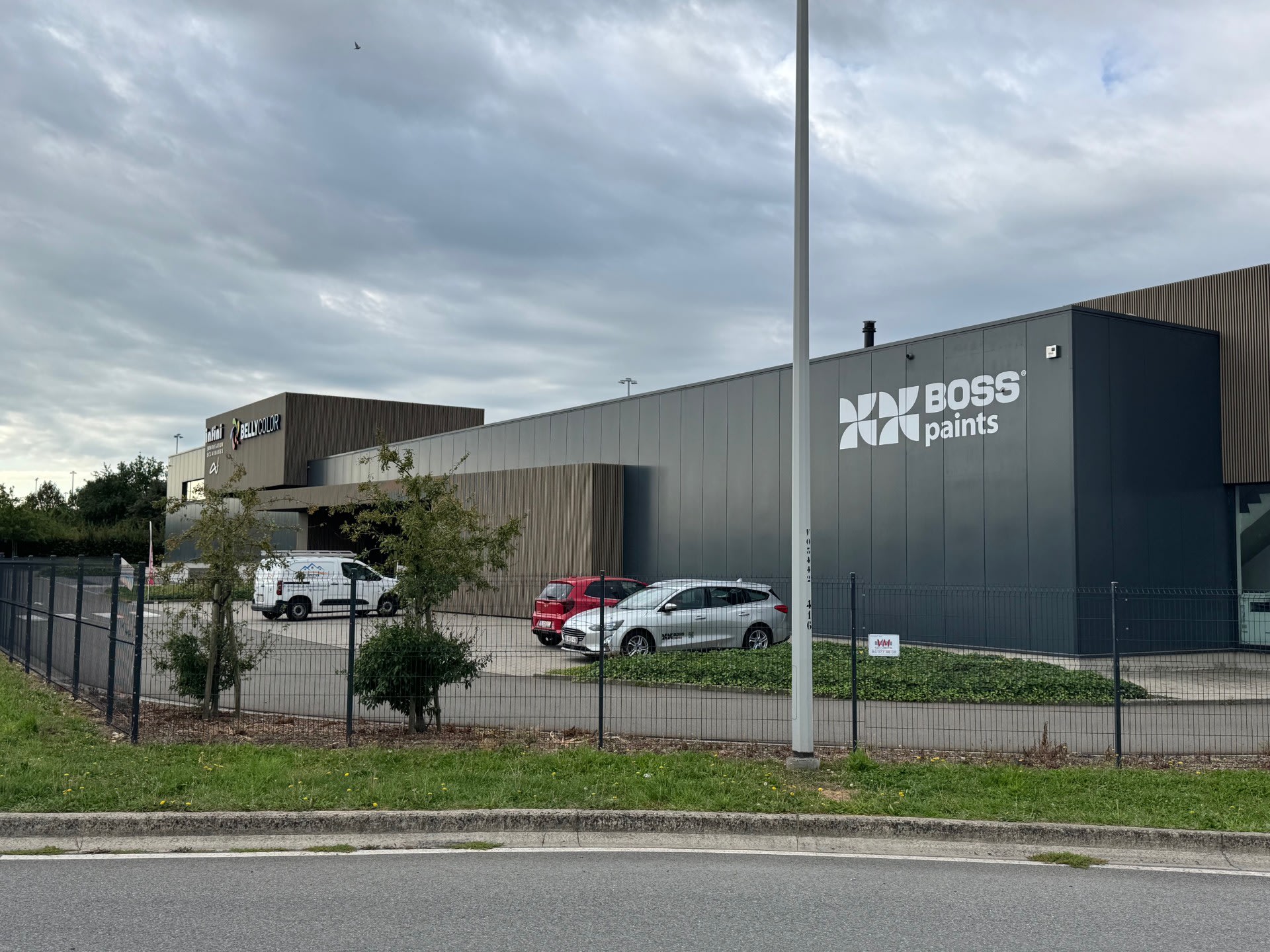 Een modern industrieel gebouw met het "BOSS paints" logo, een omheind parkeerterrein en verschillende geparkeerde voertuigen onder een bewolkte hemel.