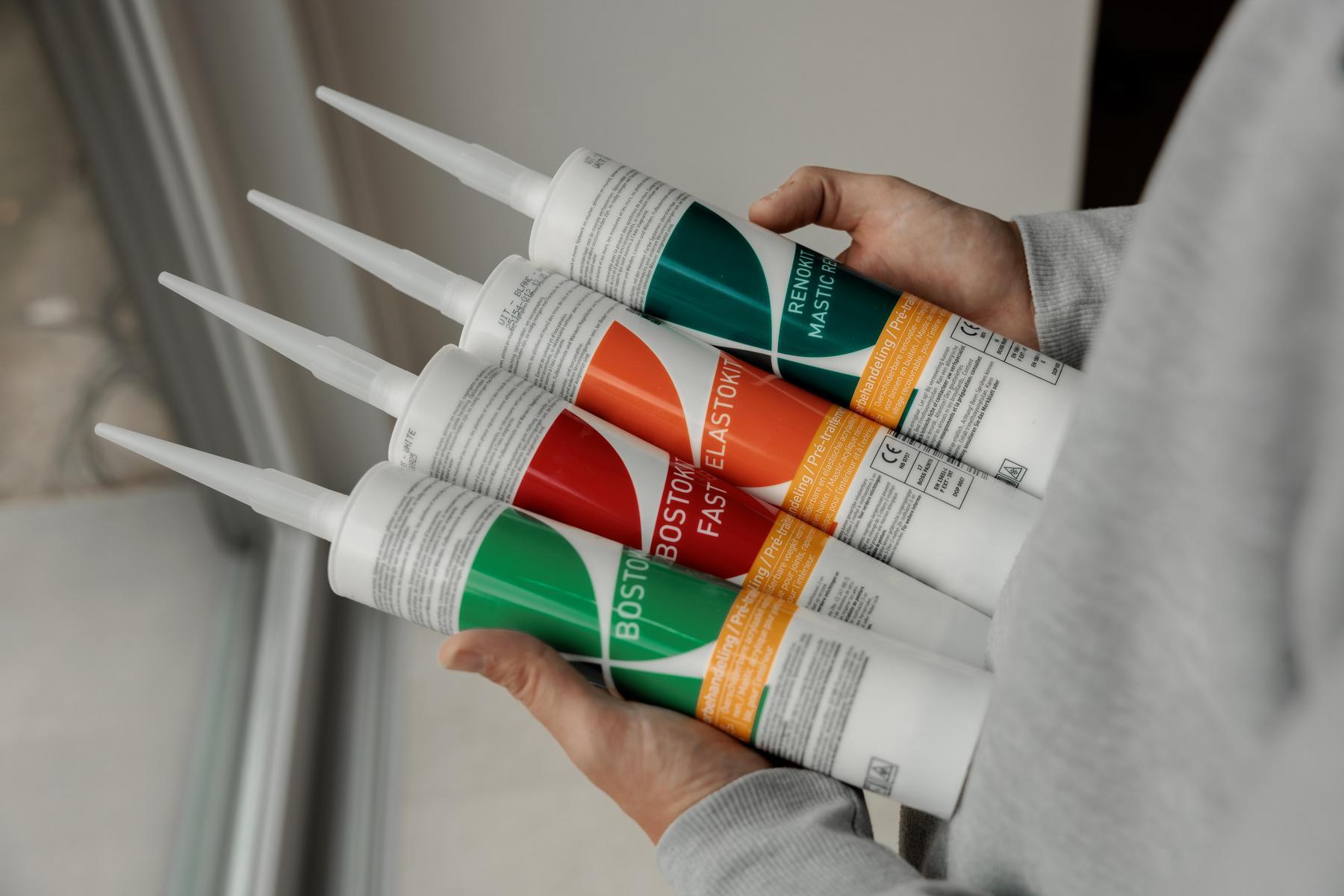 Persoon die vier tubes kit met groene, oranje en rode labels vasthoudt, van bovenaf gezien tegen een neutrale achtergrond binnenshuis.