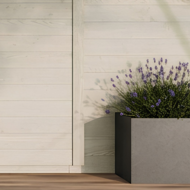 Een grijze, rechthoekige plantenbak met bloeiende lavendel staat op een houten vloer tegen een lichtgekleurde houten muur en werpt een zachte schaduw.